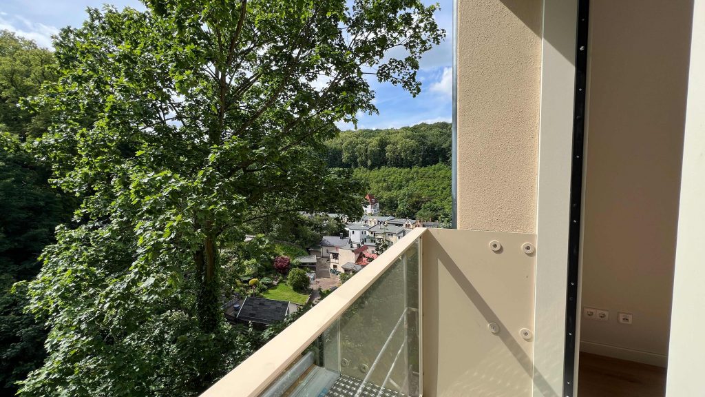 2025-06-11-15.42.34 Balkon mit Glasgeländer blickt auf bewaldete Hügel, Dorfhäuser und Kirche im Tal