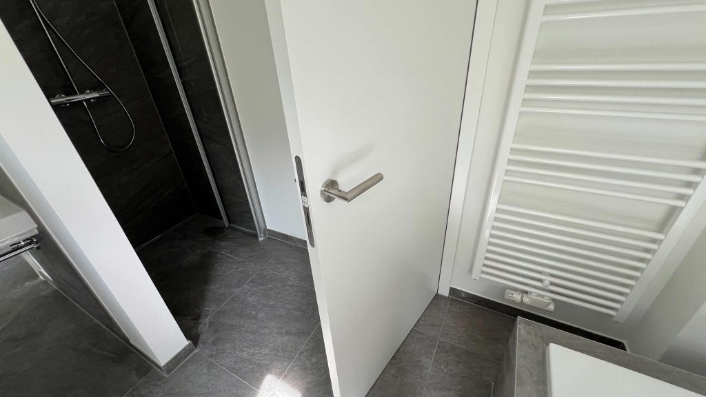 2025-06-11-15.44.13 Modernes Badezimmer mit offener Tür, dunklem Fliesenboden, weißen Wänden, Duschbereich links und Heizkörper rechts