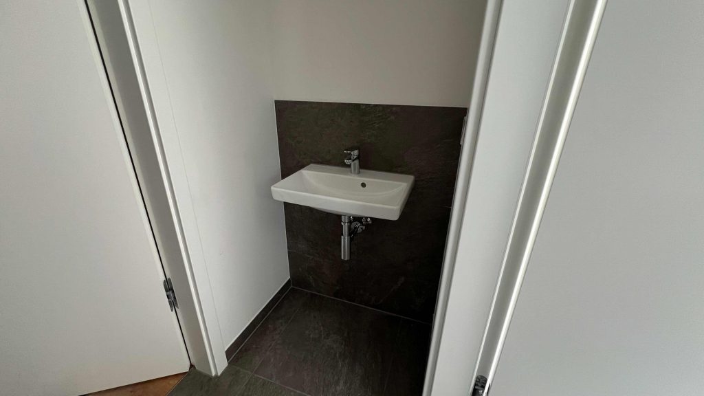 2025-06-11-15.44.58 Gästetoilette mit weißem Wandwaschbecken, schwarzer Rückwand und dunklem Fliesenboden zwischen weißen Türrahmen