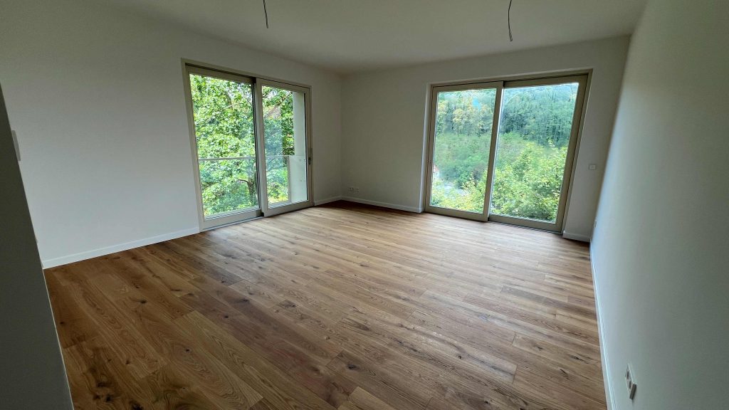 2025-07-10-12.04.39 Leeres Wohnzimmer mit Holzboden und großen Glasschiebetüren zum Waldblick, helles Tageslicht
