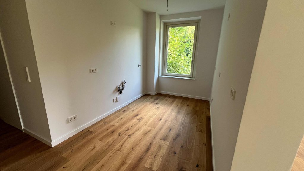 2025-07-10-12.04.42 Leeres Zimmer mit Holzboden, weißen Wänden, Fenster mit Waldblick, Steckdosen und Türöffnungen