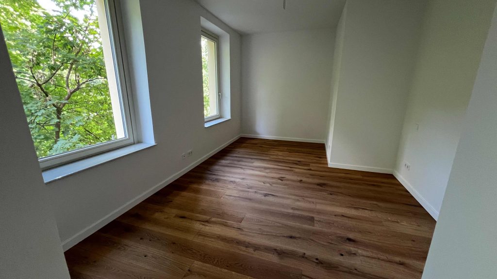 2025-07-10-12.05.40 Leeres Zimmer mit Parkettboden, hellen Wänden und zwei großen Fenstern mit Waldblick