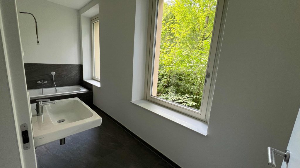 2025-07-10-12.05.46 Modernes Badezimmer mit weißem Waschbecken, schwarzer Arbeitsplatte, Badewanne im Hintergrund und großem Fenster mit Waldblick