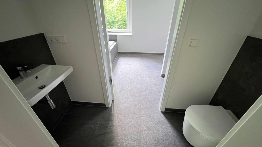 2025-07-10-12.06.58 Modernes Badezimmer mit Toilette rechts, Waschbecken links, Fliesenböden und offener Tür zum Flur mit Fensterblick