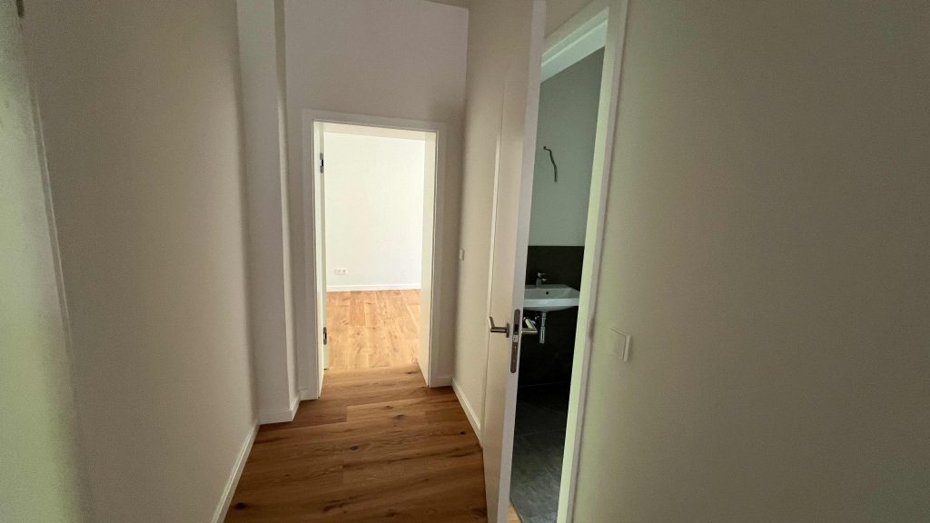 2025-07-10-12.07.15 Flur mit Holzboden, weiße Wände, Tür zum Wohnbereich links, Badezimmertür rechts mit Waschbecken sichtbar
