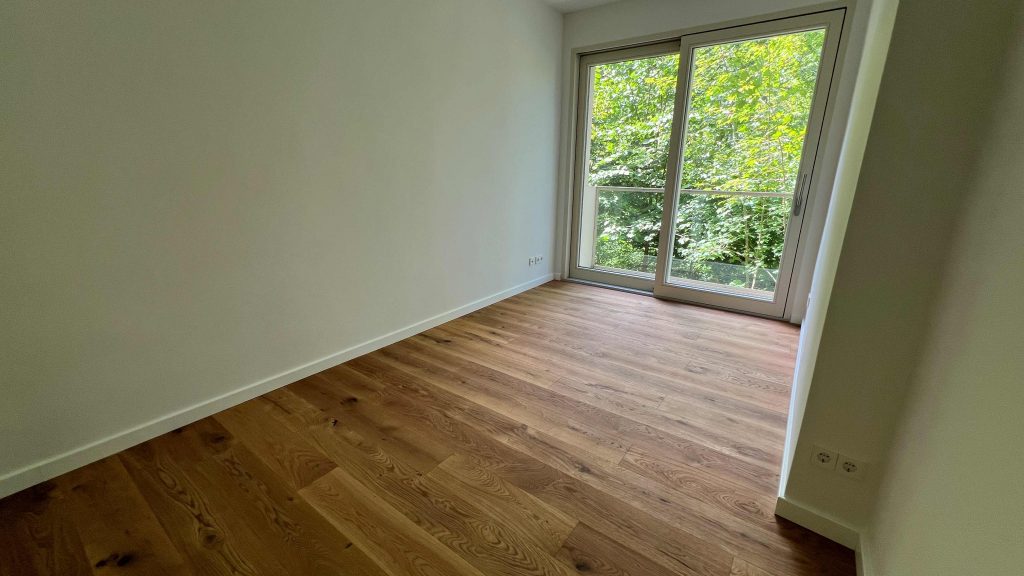 2025-07-10-12.07.23 Leeres Zimmer mit Parkettboden, hellen Wänden und großen Glastüren mit Waldblick
