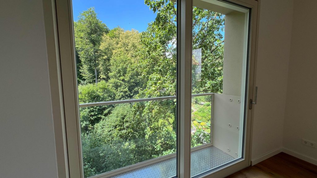 2025-07-10-12.08.10 Helles Zimmer mit offener Glasschiebetür zum Balkon, Blick auf grüne Bäume und Waldlandschaft bei sonnigem Wetter