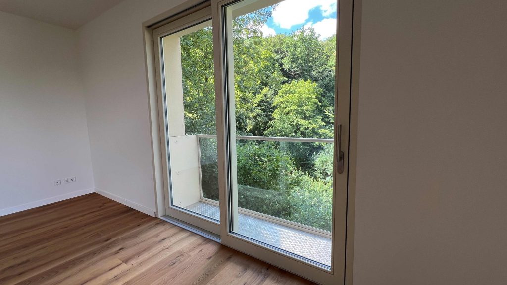 2025-07-10-12.08.18 Leeres Zimmer mit Parkett, große Glasschiebetüren zur Terrasse mit Waldblick, helle Wände, Steckdosen sichtbar