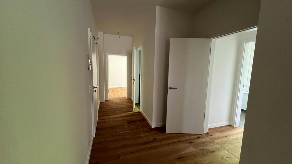 2025-07-10-12.10.58 Flur mit Holzboden, mehreren weißen Türen zu Zimmern und Badezimmer, heller Durchgang nach vorne