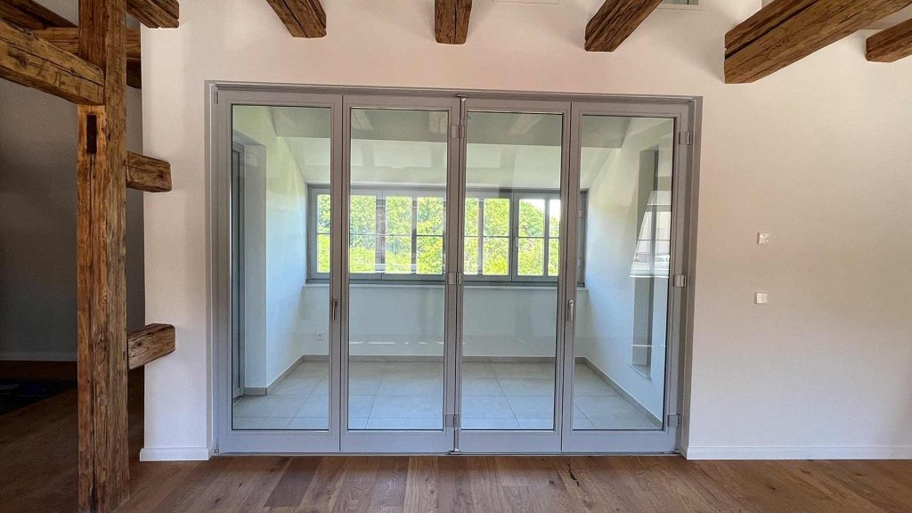 Aluminium Schiebetüren Balkon mit Holzbalken Modernes Wohnzimmer mit vier glasschiebeelementen zur Terrasse, rustikale Holzdeckenbalken, helles Tageslicht, grüne Vegetation sichtbar