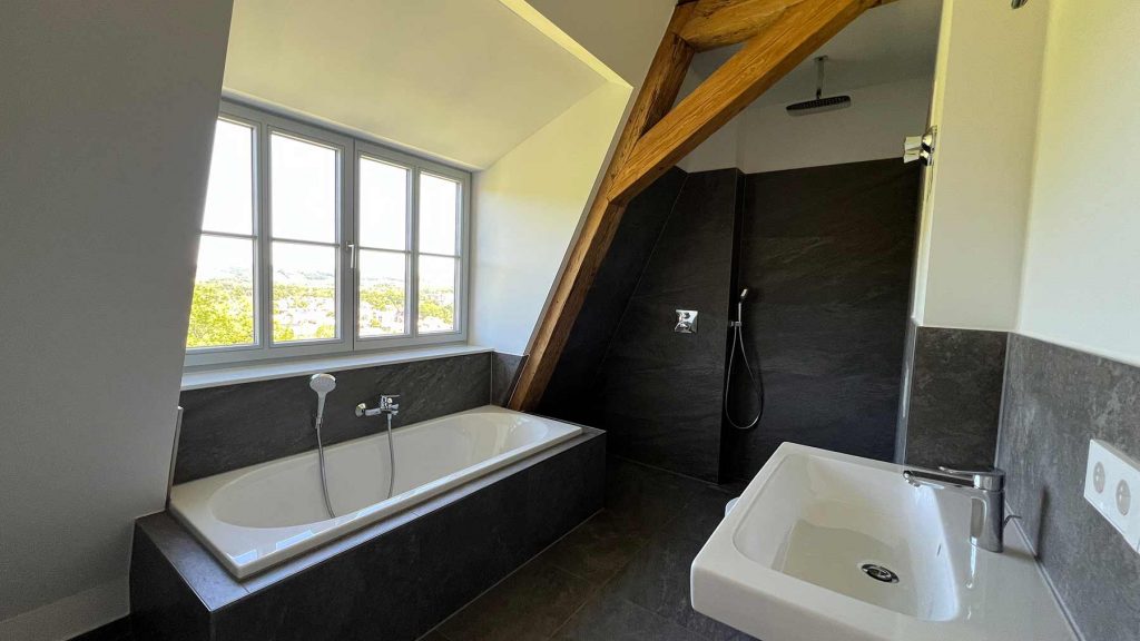 Modernes Badezimmer mit schwarzer Fliese und Holzbalken Modernes Badezimmer mit Wanne links, bodengleicher Dusche rechts, großem Fenster und Holzträgern an der Decke