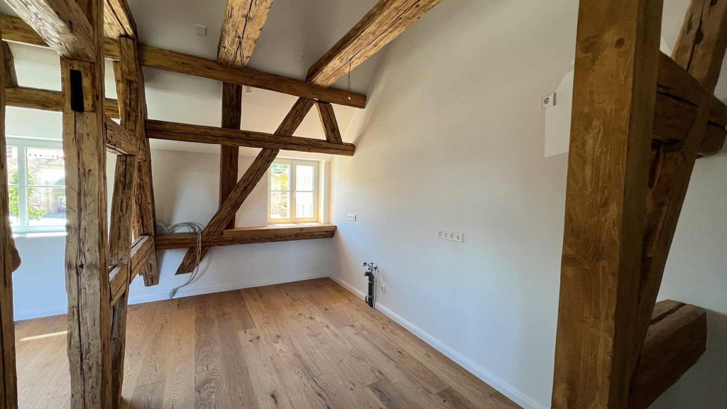 Dachgeschoss mit Holzbalken und Eichenparkett Leerer Raum mit rustikalen Holzträger an Decke und Wänden, Fenster zur Straße, Holzfußboden, weiße Wände, Steckdosen und Heizkörper