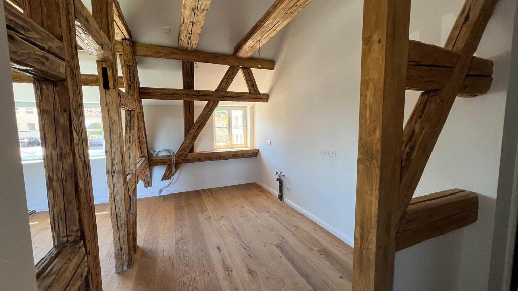 Rustikales Seitenhaus mit Holzbalken Obergeschoss Leeres Obergeschoss mit massiven Holzträgern, Parkettboden, großem Fenster und offener Tür zum Treppenhaus
