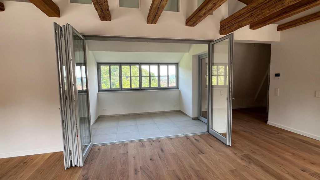 Modernes Wohnzimmer mit Glasschiebetüren und Holzbalken Geräumiges Wohnzimmer mit großflächigen Glasschiebetüren zur Terrasse, Holzdeckenbalken, Parkettboden und angrenzender Küche