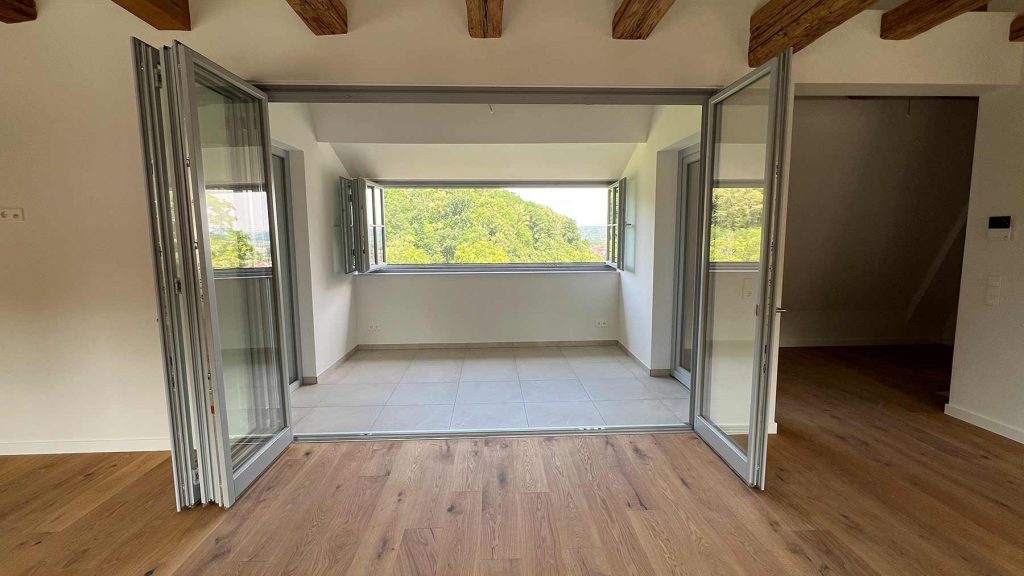 Moderne Falttüren Terrasse Holzboden Ausblick Wohnraum mit geöffneten Glasschiebetüren zur Terrasse, Blick auf grüne Landschaft, Holzparkettboden, weiße Wände, Dachbalken