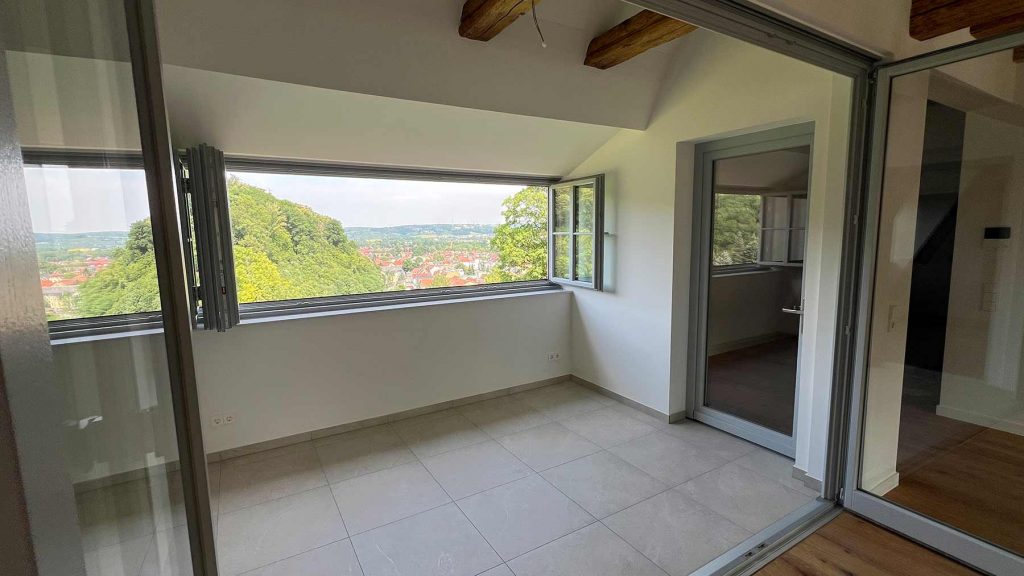 Panorama-Terrasse mit Panoramafenstern Landschaftsblick Helles Wohnzimmer mit großflächigen Fenstern, Blick auf bewaldete Hügellandschaft und Dorf, Glastüren zum Nebenraum, Holzdeckenbalken, Fliesenboden