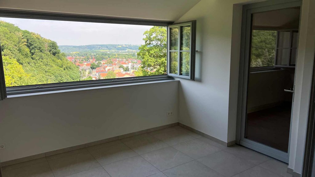 Modernes Wohnzimmer mit Panoramafenster Bergblick Leeres Wohnzimmer mit Panoramafenstern, Blick auf Stadt mit roten Dächern und grüne Hügel, Glastür rechts, helle Fliesen