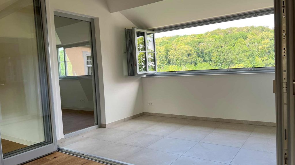 Moderne Wohnfläche mit Panoramafenstern und Waldblick Leeres Wohnzimmer mit Fliesenboden, Glasschiebetür links, großes Fenster mit Waldaussicht rechts, weiße Wände, moderne Architektur