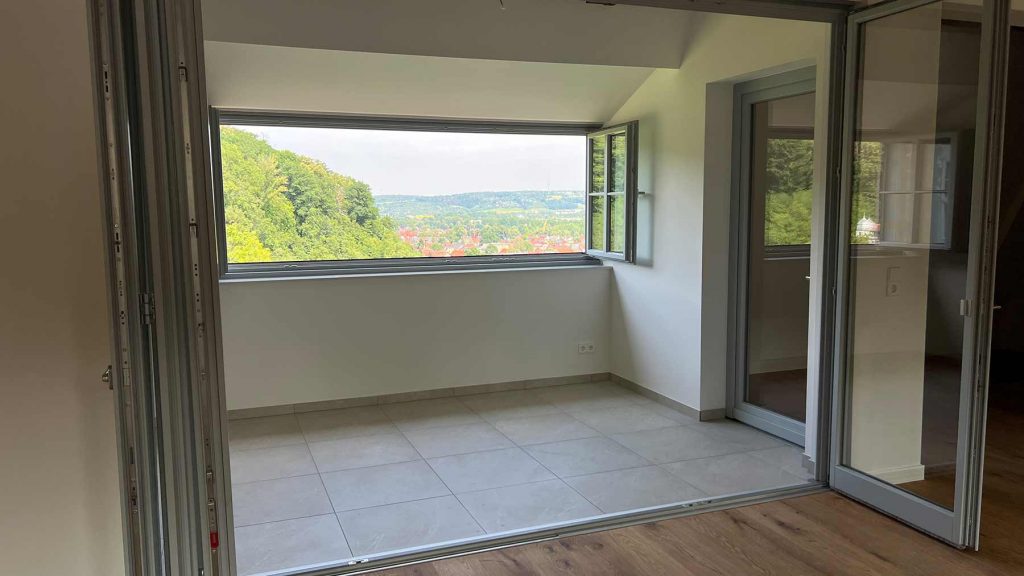Modernes Wohnzimmer mit Panoramafenster Waldblick Leeres Wohnzimmer mit großflächigem Fenster, Glastüren zur Terrasse, Ausblick auf bewaldete Hügellandschaft mit roten Dachziegeln