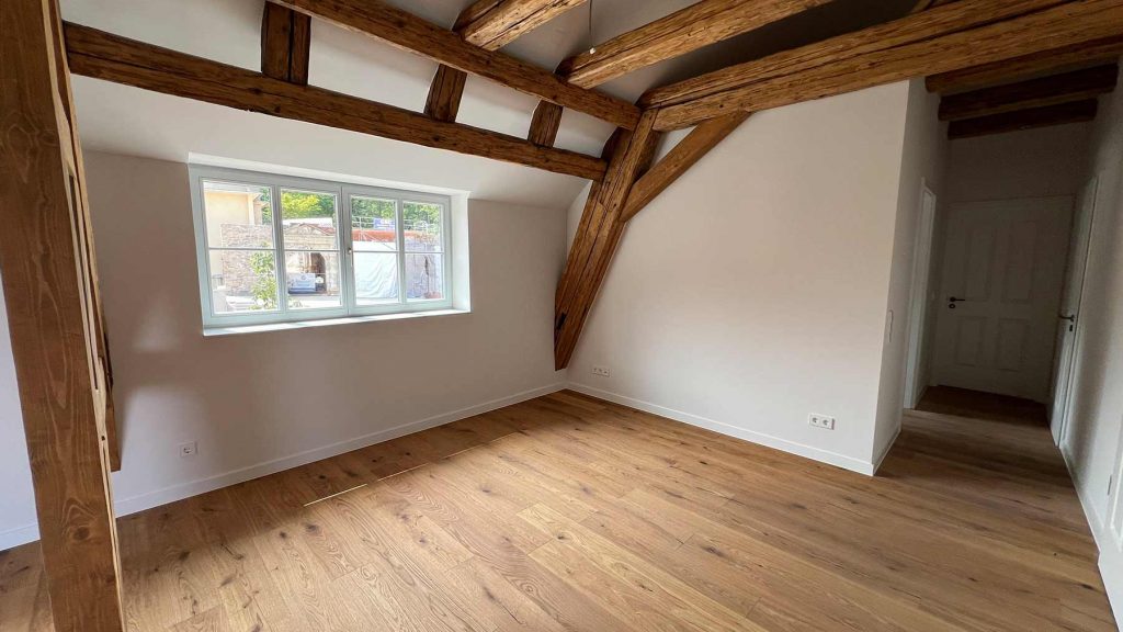 Dachgeschoss Raum mit Holzbalken und Eichenparkett Leeres Wohnzimmer im Obergeschoss mit exponierten Holzbalken, großem Fenster zur Straße und Durchgang zu angrenzenden Räumen