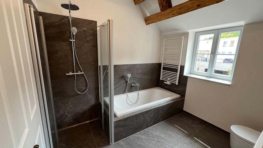 Modernes Badezimmer mit Dusche und Badewanne Modernes Badezimmer mit begehbarer Duschkabine links, Badewanne mit Duschaufsatz rechts, Handtuchheizkörper, Fenster mit Außenblick, Holzbalken an der Decke, helle Fliesen und dunkle Akzente