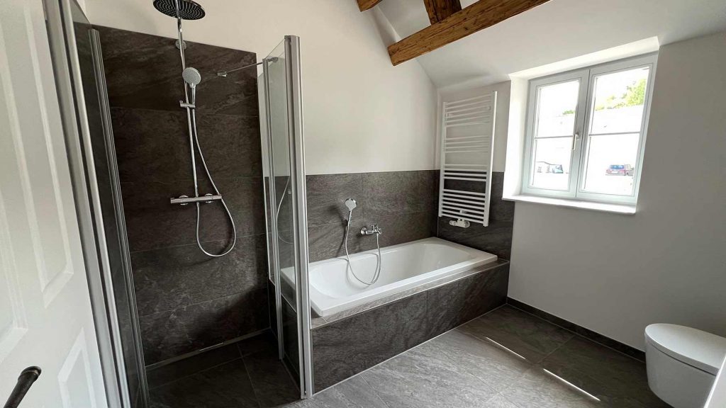 Modernes Badezimmer Dusche Wanne Anthrazit Geräumiges Badezimmer mit separater Duschkabine links, Badewanne mit Handbrause rechts, dunklen Fliesen, Holzbalken an der Decke, großem Fenster und WC
