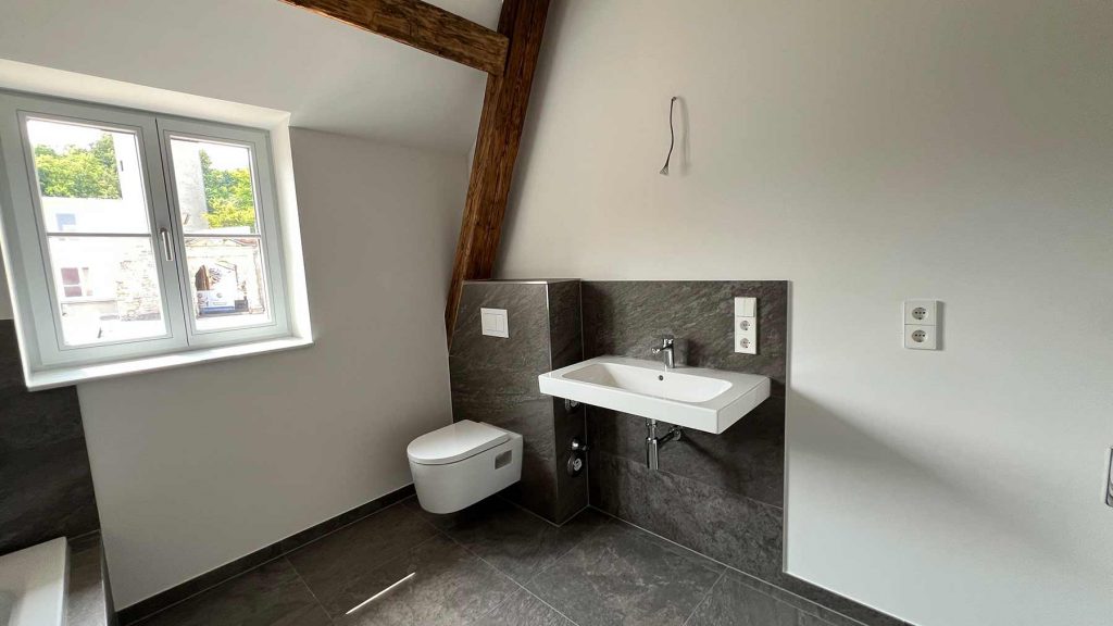 Modernes Badezimmer Grau-Weiß mit Wandtoilette Helles Badezimmer mit Wandklosett links, rechteckigem Waschbecken auf dunkler Steinplatte, Doppelfenster, Holzbalken und Steckdosen