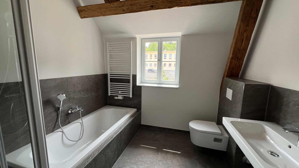 Modernes Badezimmer Grau-Weiß Dachgeschoss Badezimmer mit Badewanne links, Toilette und Doppelwaschbecken rechts, Fenster zur Straße, dunkle Bodenfliesen, Holzbalken an der Decke