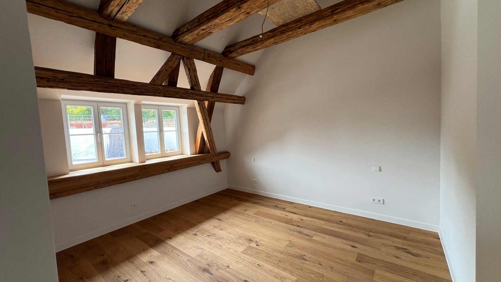 Dachzimmer Holzbalken Dielenboden Obergeschoss Leeres Dachzimmer mit Fensterfront, massiven Holzbalken an Decke und Wänden, Parkettboden, weiße Wandflächen, moderne Elektroinstallationen