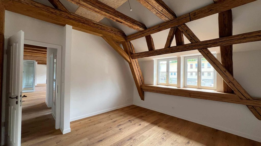Dachgeschoss Loft Holzbalken Wohnraum Modern Dachgeschoss-Zimmer mit Holzdeckenbalken, Fenster zur Straße, Holzfußboden, weiße Wände, offene Tür zum Flur
