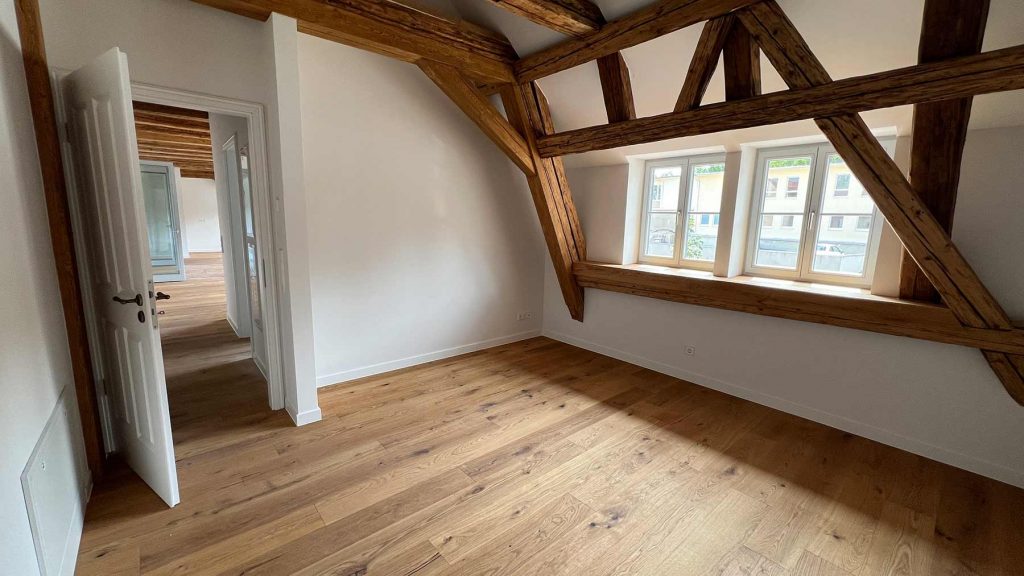 Dachgeschoss Wohnzimmer Holzbalken Parkettboden hell Helles Dachzimmer mit Holzbalkendecke, Fensterfront zur Straße, weißer Tür links, offene Verbindung zu weiteren Räumen