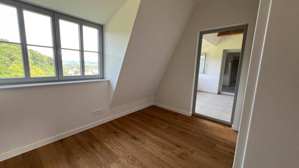 Dachgeschoss Zimmer Holzboden Panoramafenster Wohnraum Leeres Dachzimmer mit Parkettboden, Schrägwand, großem Fenster mit Landschaftsblick und offener Türe zum angrenzenden Raum