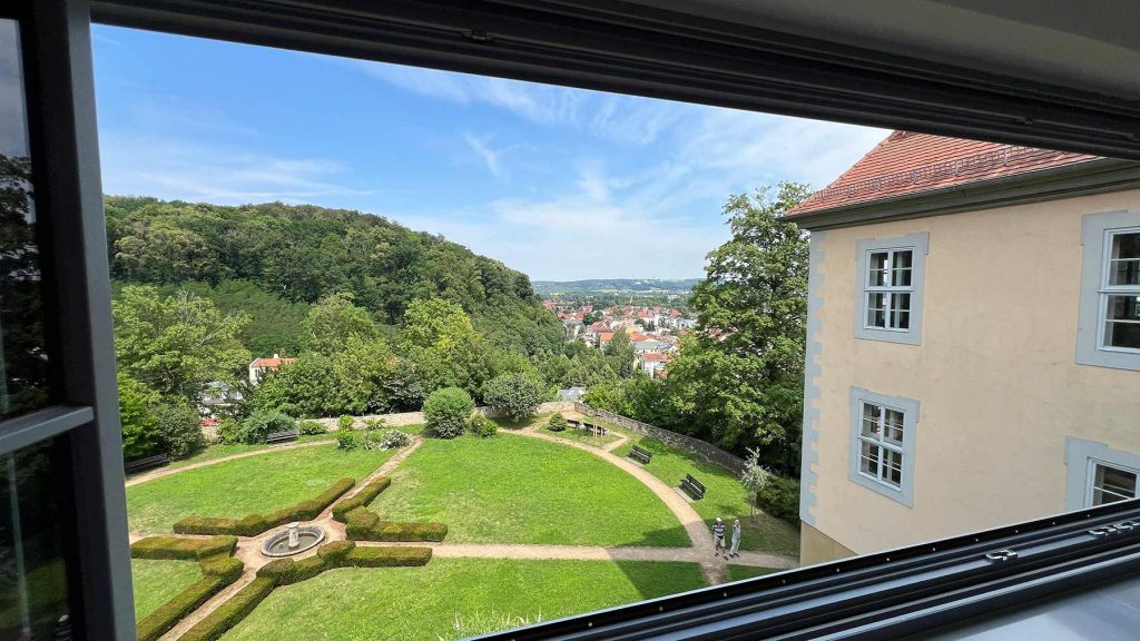Herrschaftliche Gartenanlage Schloss Panoramablick Stadt Blick aus Fenster auf gepflegten Rasen mit Weganlage, Brunnen, Bäume und Stadt im Hintergrund