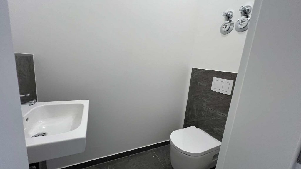 Modernes Badezimmer Weiß Anthrazit Fliesen Modernes WC mit Spülplatte an anthrazitfarbiger Wand, weißem Waschbecken links, Handtuchhalter und zwei Wandleuchten oben rechts