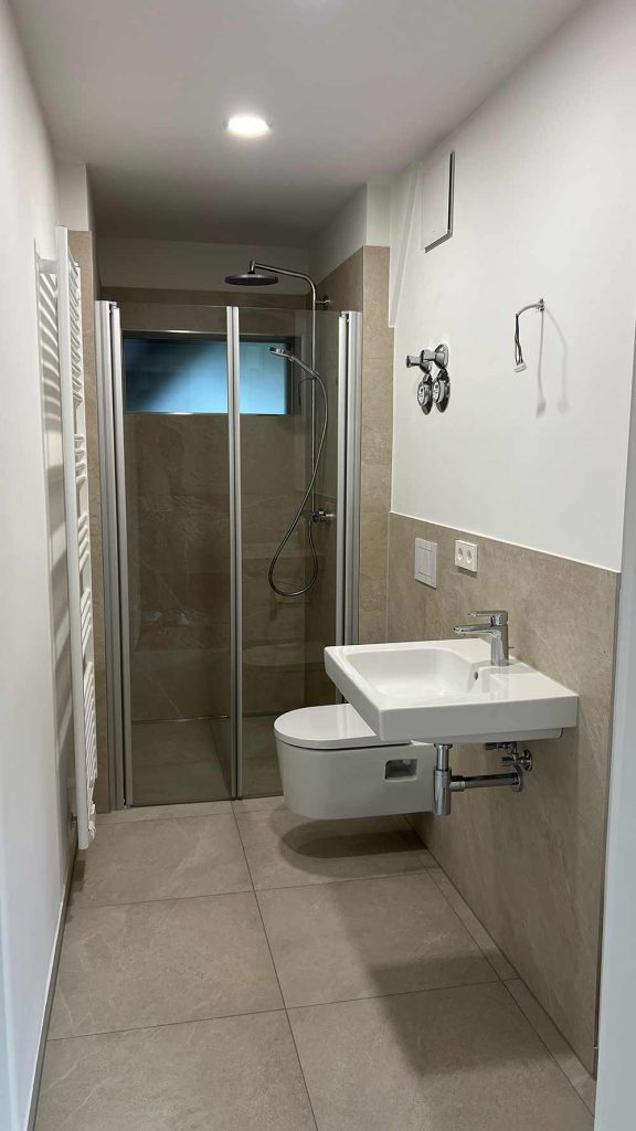 Modernes Badezimmer mit Dusche und Waschbecken Schmales Badezimmer mit Toilette links, Waschbecken rechts, Duschkabine mit Glastüren hinten, Handtuchhalter, beige Bodenfliesen