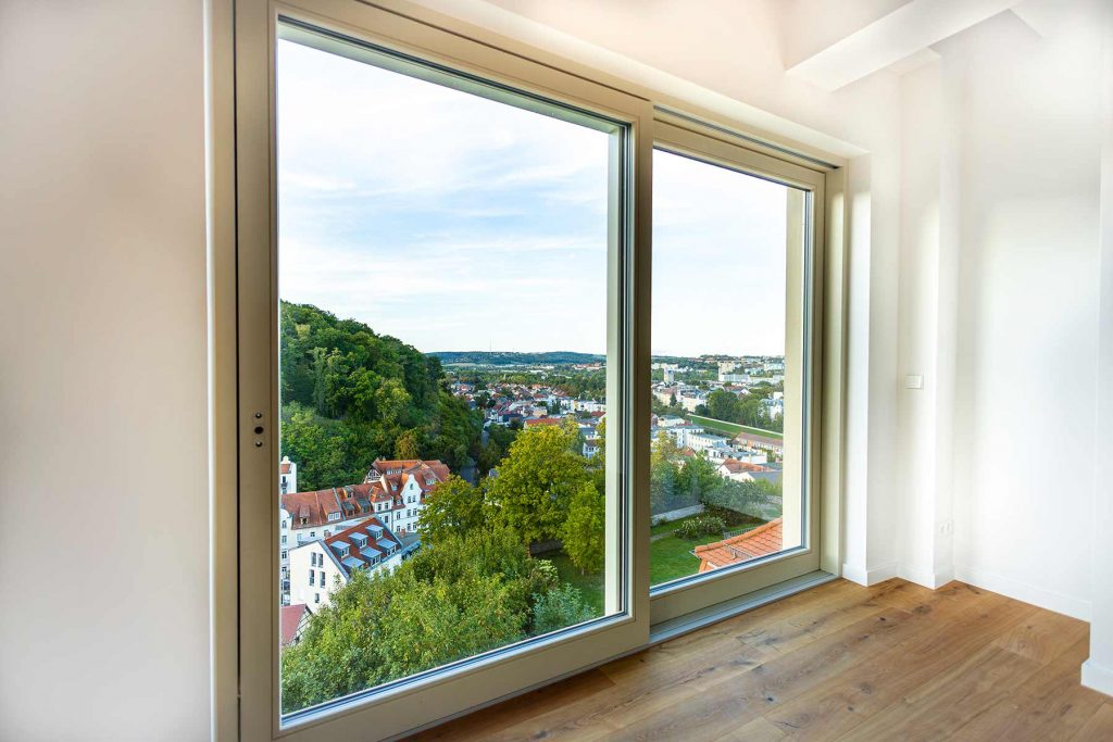 7I4A4990-2 Große Glasfenster mit Blick auf bewaldete Hügel, historische Häuser mit Ziegeldächern und Stadtlandschaft, moderner Holzboden und weiße Wände