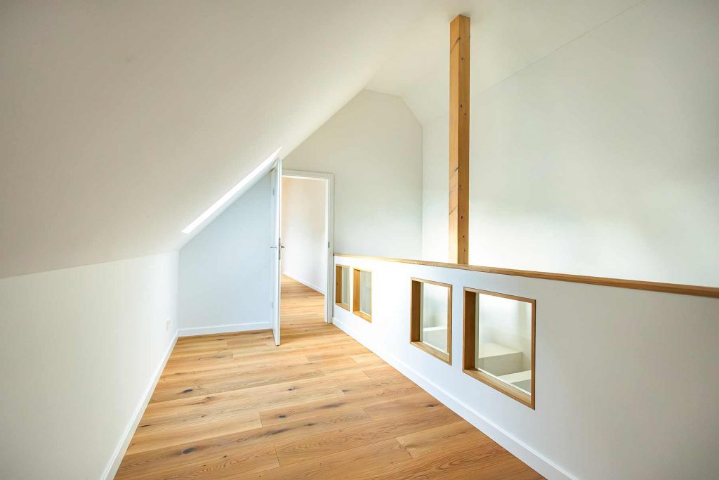 7I4A5022 Dachgeschoss-Flur mit Holzdielen, weißen Wänden, Holzbalken, offener Tür zu Nebenraum und Fensterausblicken