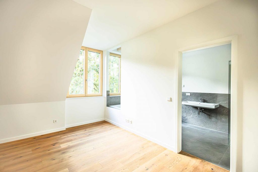 7I4A5087 Helles Dachgeschosszimmer mit Holzboden, großem Fenster zu Baumblick, offene Badezimmertür mit grauer Fliesen und Waschbecken sichtbar
