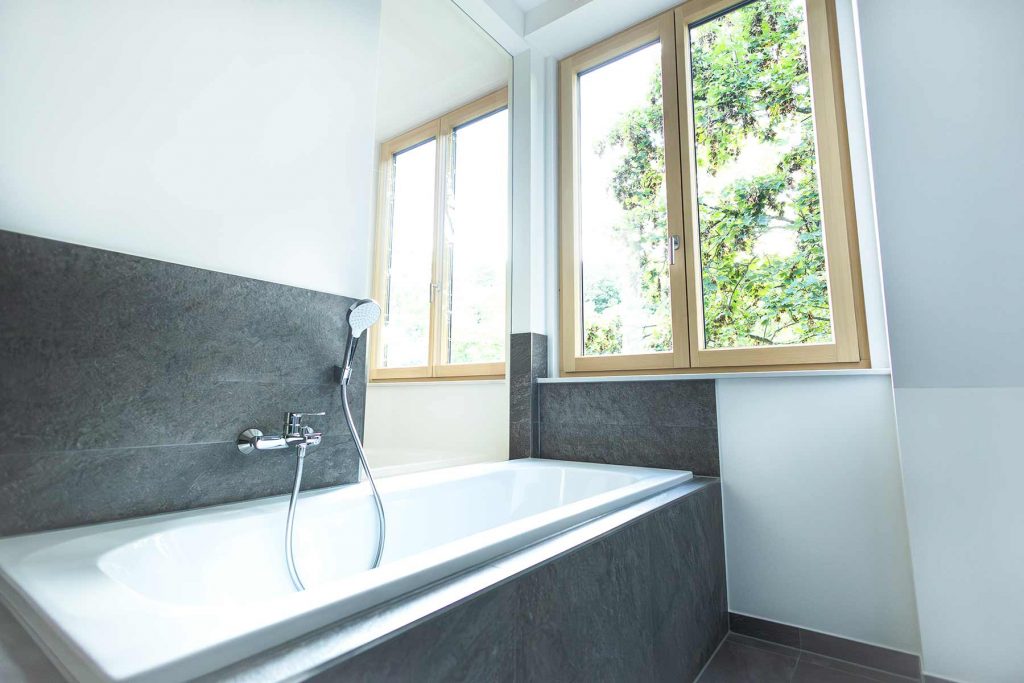 7I4A5109 Modernes Badezimmer mit Badewanne, grauer Fliesenverkleidung, Handbrause und großen Fenstern mit Waldblick