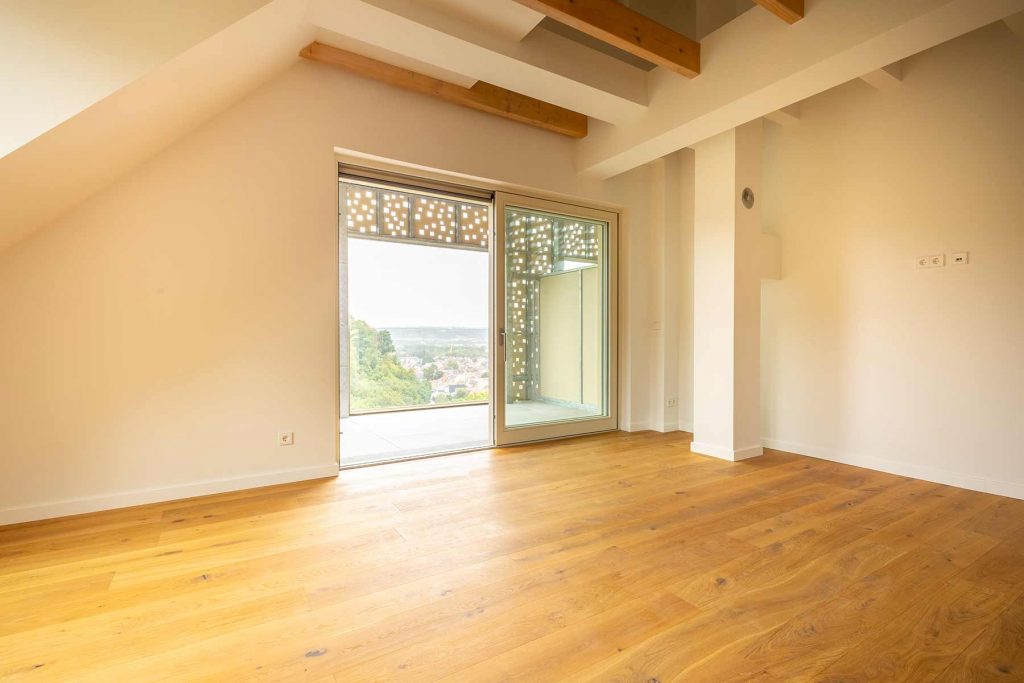 Modernes Wohnzimmer Holzboden Dachgeschoss Waldblick Leeres Wohnzimmer mit Parkettboden, Dachbalken, großer Terrassentür mit Sonnenschutz und Ausblick auf grüne Landschaft