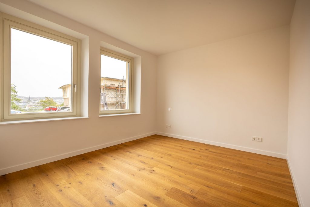 Leeres Zimmer Holzboden Fenster Stadtblick Helles Wohnzimmer mit Parkettboden, zwei großen Fenstern zur Stadt, beige Wände und Steckdosen