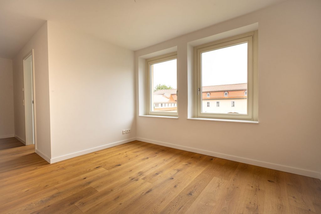 Leeres Wohnzimmer Parkett Fenster Stadtblick Helles Wohnzimmer mit Holzboden, zwei großen Fenstern mit Blick auf orange-weißes Gebäude, weiße Wände, offene Tür links, Steckdosen