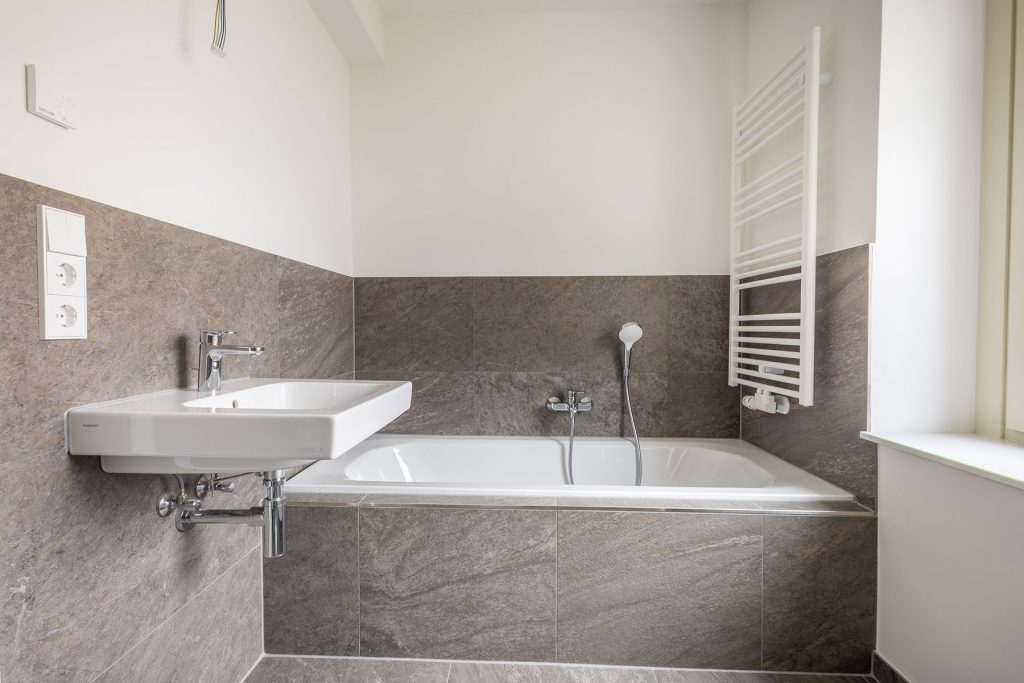 Modernes Badezimmer Grau-Weiß mit Wanne Modernes Badezimmer mit Wanne, Waschbecken links an grauer Fliesenwand, weiße Handtuchwärmer rechts, helles Fenster