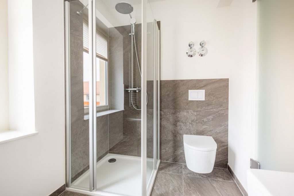 Modernes Badezimmer Graue Fliesen Dusche Toilette Helles Bad mit grauem Steindekor, bodenebener Glasdusche, weißer Toilette, Fenster und Armatur mit Regenduschkopf