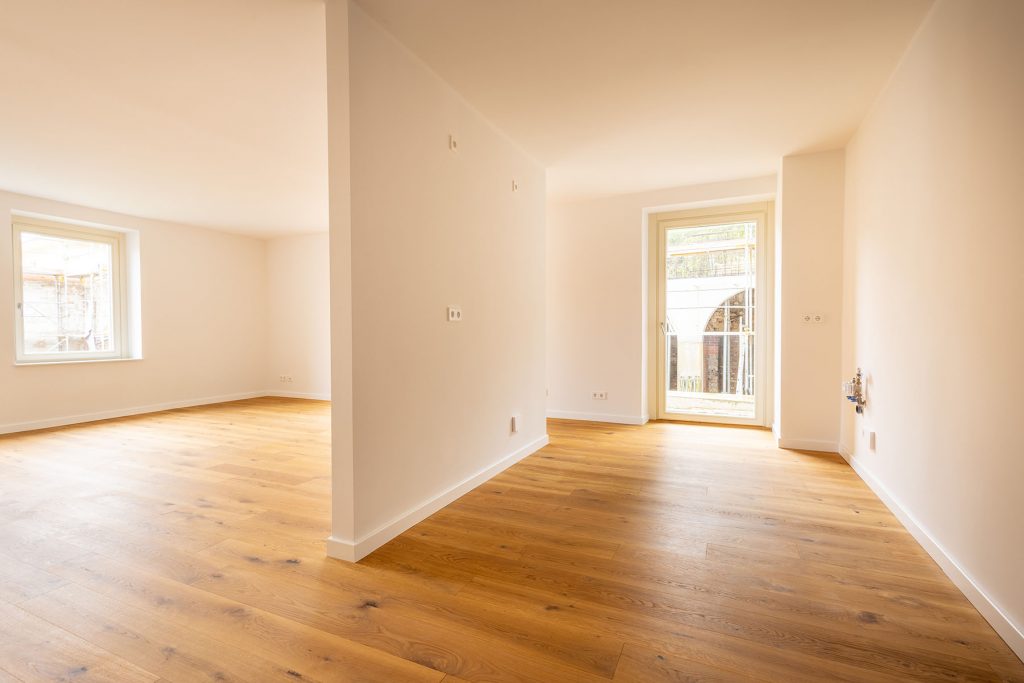 Helles Wohnzimmer Eichenparkett Balkonzugang Geräumiges Wohnzimmer mit Holzböden, hellen Wänden, zwei Fenstern und Glastür zum Balkon, offene Raumaufteilung mit Säulen