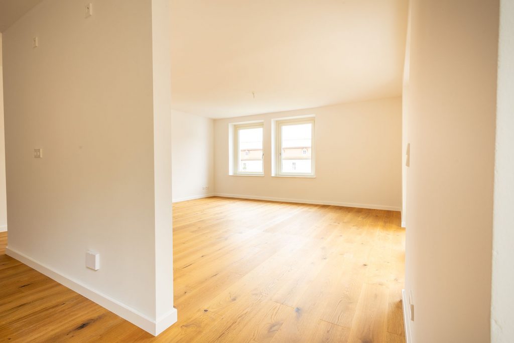 Leeres Wohnzimmer Parkett Fenster Neubau Helles Wohnzimmer mit Holzboden, weißen Wänden, zwei Fenstern und offener Raumaufteilung mit Säulen