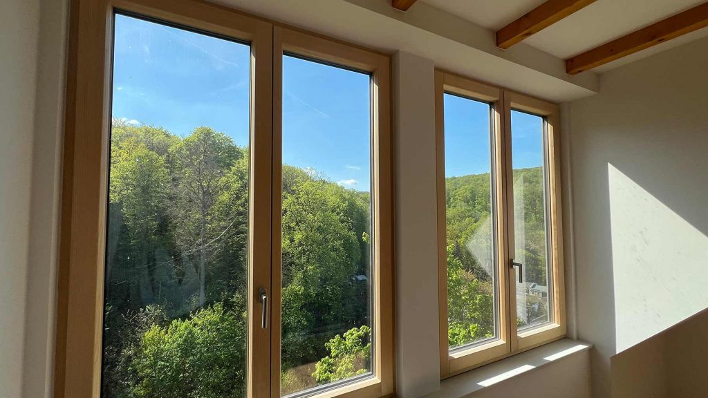 WE-17-2025-04-30-15.29.06 Dachgeschossraum mit vier Holzfenstern, Blick auf bewaldete Hügellandschaft unter blauem Himmel, Sonnenlicht fällt auf helle Wand